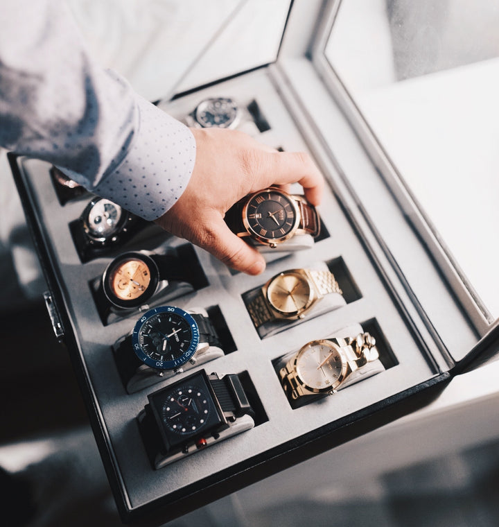 man reaching to pick up a watch from 10 luxury watches in a watch case.